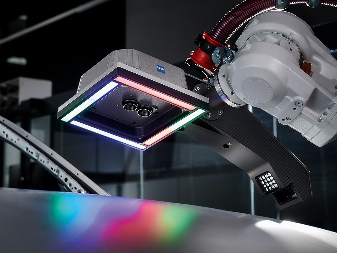 Ein mit einem ZEISS 3D-Scansensor ausgestatteter Roboterarm schwebt über einer Metalloberfläche, die von mehrfarbigen LED-Leuchten beleuchtet wird.