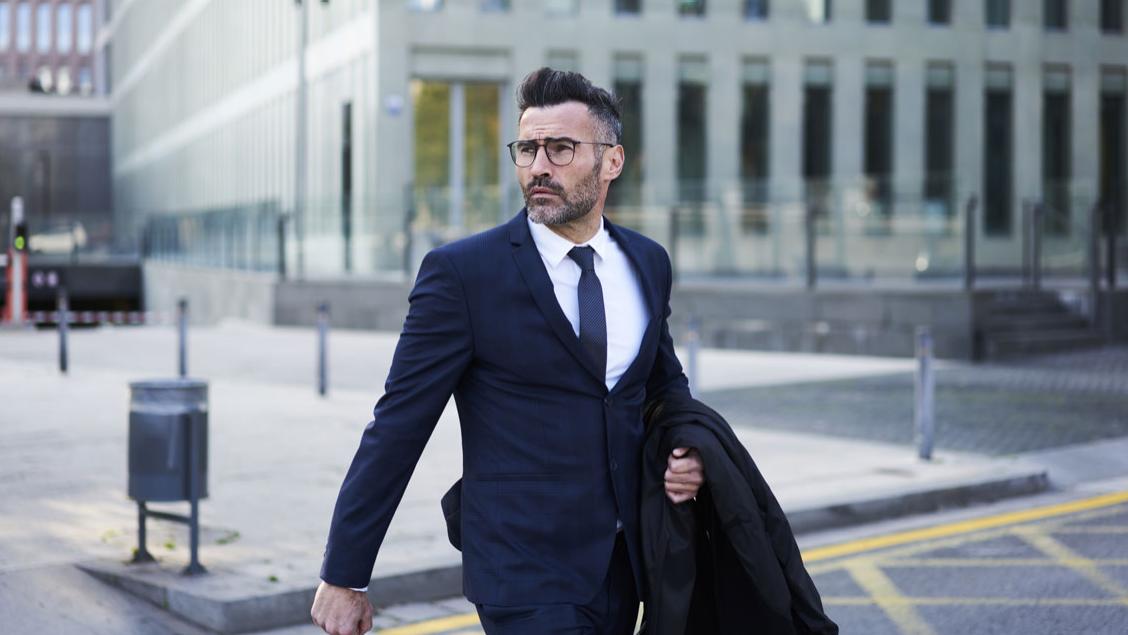 Confident handsome bearded male lawyer in trendy eyeglasses crossing street passing modern constructed architecture buildings with headquarters in business district of city hurrying for meeting.